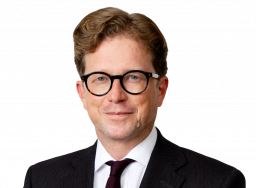 A man with wavy brown hair and glasses, wearing a dark suit, white shirt, burgundy tie, and patterned pocket square, poses against a plain light background—an image capturing Noel Casey KC.
