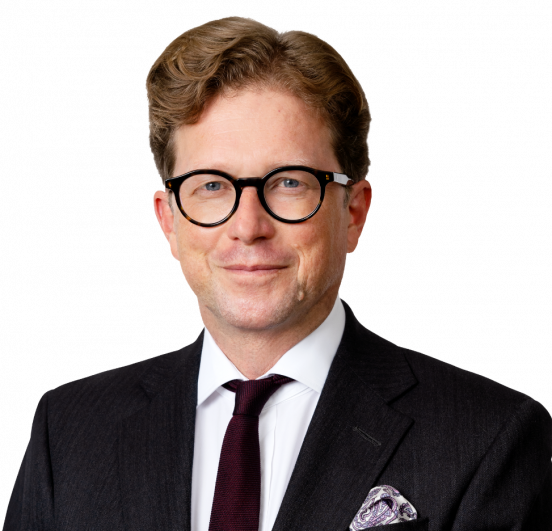 A man with wavy brown hair and glasses, wearing a dark suit, white shirt, burgundy tie, and patterned pocket square, poses against a plain light background—an image capturing Noel Casey KC.