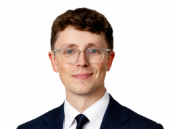 A man with short brown hair and glasses in a dark suit and tie.