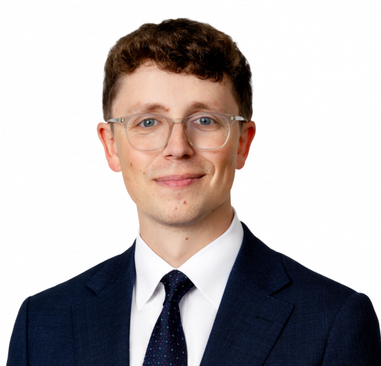 A man with short brown hair and glasses in a dark suit and tie.